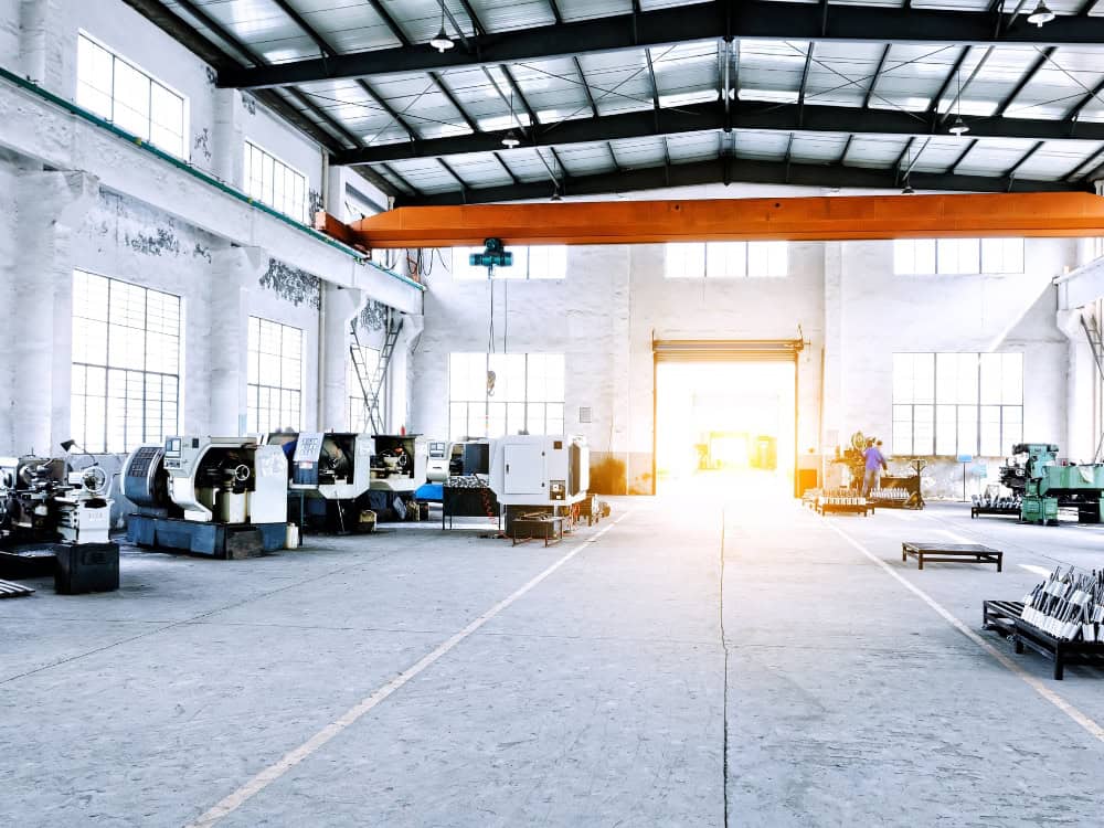 A large, empty factory with rows of machinery. Sunlight streams in through a large doorway. Uneven floors are visible.